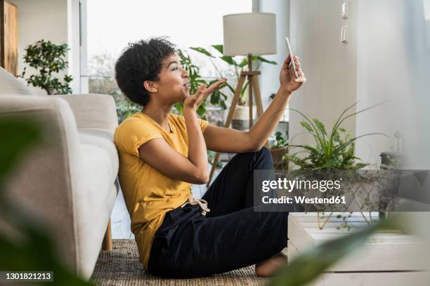 woman blowing a kiss on video call through mobile phone while sitting by sofa at home - mandare un bacio foto e immagini stock