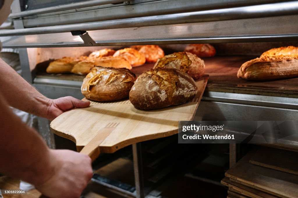 Male chef with pizza peel removing baked bread from oven at bakery