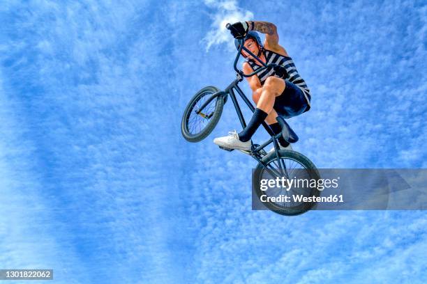 young man performing stunt with bicycle against blue sky - bmx stock-fotos und bilder