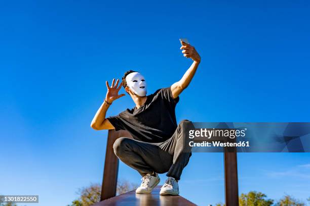 young man wearing white mask waving hand to video call through mobile phone while crouching on retaining wall - attore foto e immagini stock