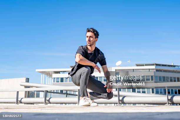 man looking away while crouching on rooftop against sky - hombre agachado fotografías e imágenes de stock