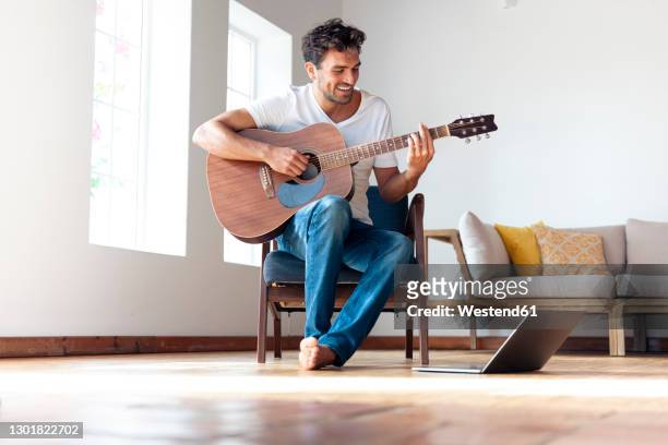 mid adult man practicing guitar while sitting on chair by laptop at home - guitarrista fotografías e imágenes de stock