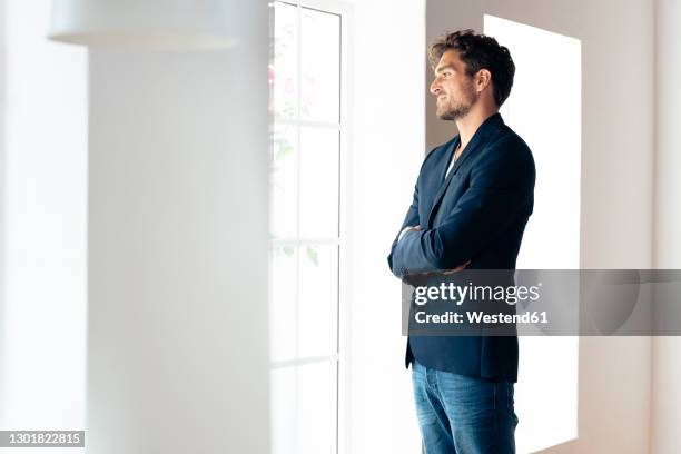 smiling businessman looking through window while standing with arms crossed at home - man side standing stock-fotos und bilder