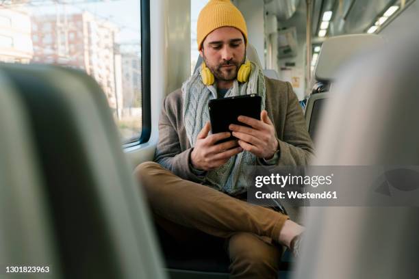 man wearing knit hat and headphone using digital tablet while sitting in train - mid adult fotografías e imágenes de stock