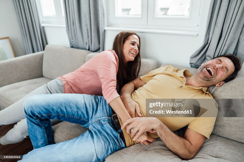 Happy mature woman tickling man while sitting on sofa at home