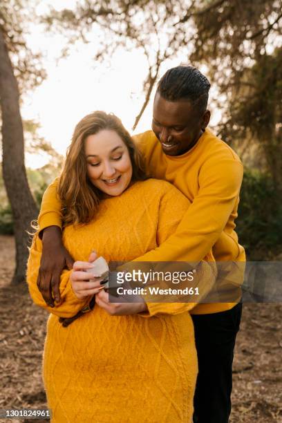 man embracing woman smiling while holding engagement ring box at forest - black women engagement rings foto e immagini stock