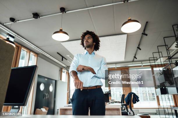 confident young male professional rolling up sleeves in office - rolling up sleeve stock-fotos und bilder