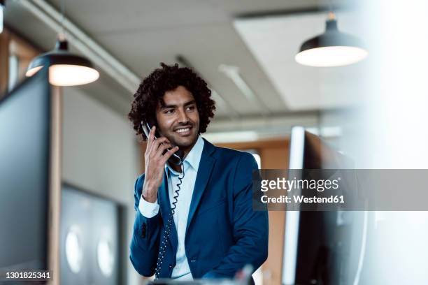 smiling young businessman talking on telephone while looking away at workplace - festnetztelefon stock-fotos und bilder