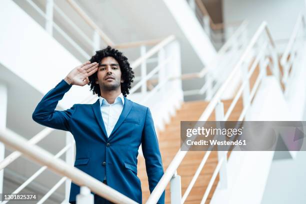 confident young male professional saluting while standing at staircase in office - saluting stock-fotos und bilder