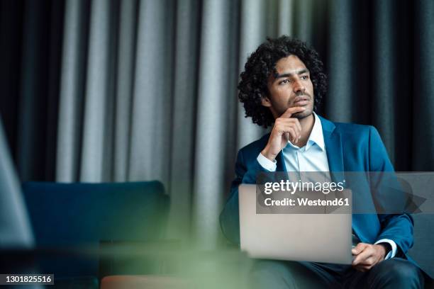 thoughtful male entrepreneur looking away while sitting with laptop at office - hand on chin stock pictures, royalty-free photos & images