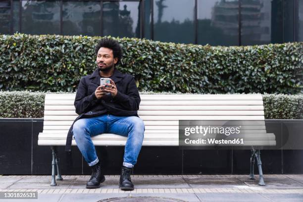 mid adult man with afro hair using smart phone while sitting on bench in city - man daydreaming stock pictures, royalty-free photos & images