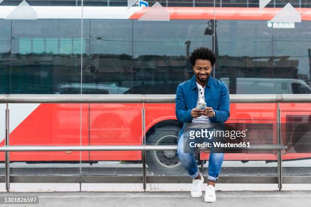 smiling stylish mid adult man using mobile phone while sitting at bus stop - aspettare foto e immagini stock