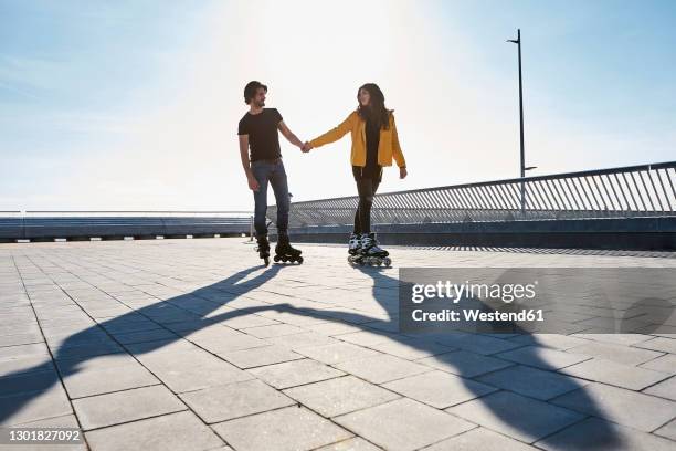 young couple looking at each other while roller skating on pier during sunny day - long shadow stock pictures, royalty-free photos & images