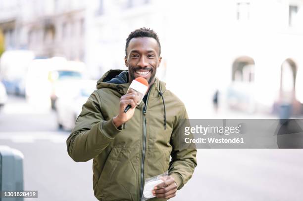 smiling male journalist with microphone standing on street in city - news anchor stock-fotos und bilder