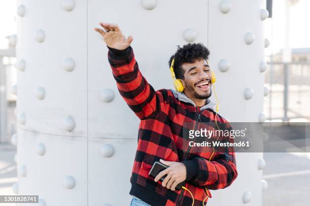 happy man with mobile phone listening music through headphones while dancing against wall - young man listening to music on smart phone outdoors stockfoto's en -beelden
