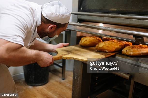 male baker removing breads from oven in kitchen at bakery - bakning business bildbanksfoton och bilder