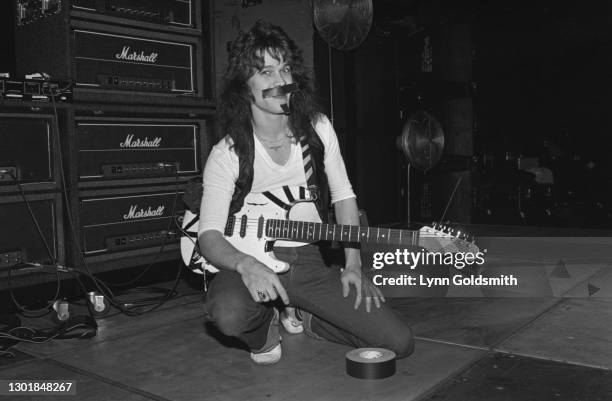 American guitarist and songwriter Eddie Van Halen wearing a white t-shirt beneath a waistcoat, with a moustache and soul patch made from gaffa tape,...