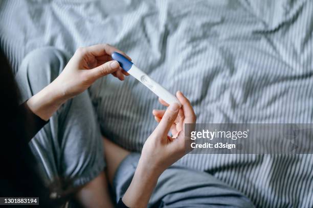 high angle view of disappointed young asian woman sitting on the bed and holding a negative pregnancy test. life events, infertility and family concept - infertilidad fotografías e imágenes de stock
