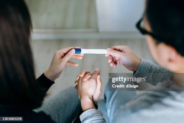 high angle view of an affectionate young asian couple sitting on the bed, holding hands and holding a positive pregnancy test together. it's finally happening. the long-awaited news. life events, fertility and family concept - infertility stock pictures, royalty-free photos & images