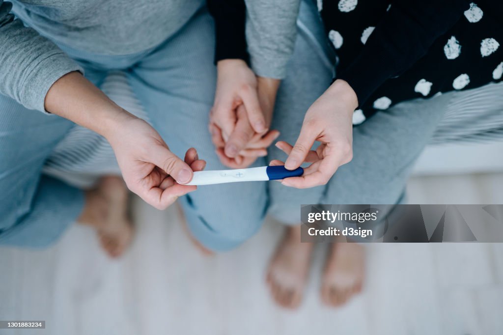 Overhead view of an affectionate young Asian couple sitting on the bed, holding hands and holding a positive pregnancy test together. It's finally happening. The long-awaited news. Life events, fertility and family concept