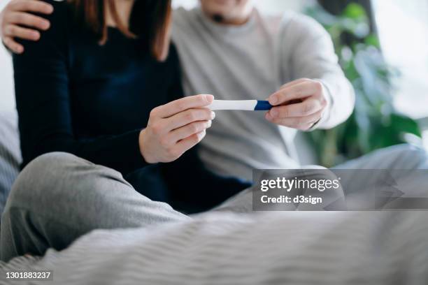 cropped shot of an affectionate young asian couple sitting on the bed holding a pregnancy test together, waiting for the result of the test. life events, fertility and family concept - gynecological examination stock-fotos und bilder
