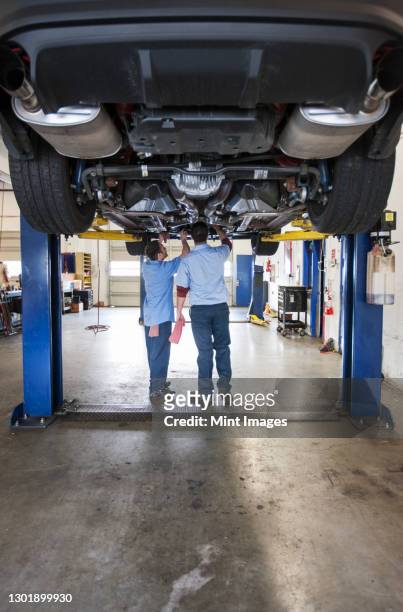 two mechanics work on the underside of a care on a lift in a repair shop - hydraulics stock-fotos und bilder