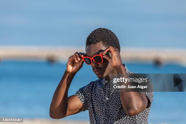 confident black man putting on sunglasses on beach - aufsetzen stock-fotos und bilder