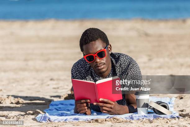 trendy ethnic man reading book on sandy beach - beach book reading stock-fotos und bilder