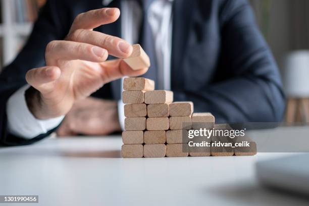 businessman hand pulling out or placing wood block on the tower and semi-pyramid in modern office. plan and strategy in business. - stability stock pictures, royalty-free photos & images