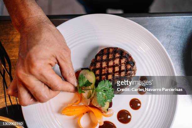 hand of a chef garnishing a fillet of beef with herbs and vegetables on a ceramic plate in a kitchen - dining experience stock pictures, royalty-free photos & images