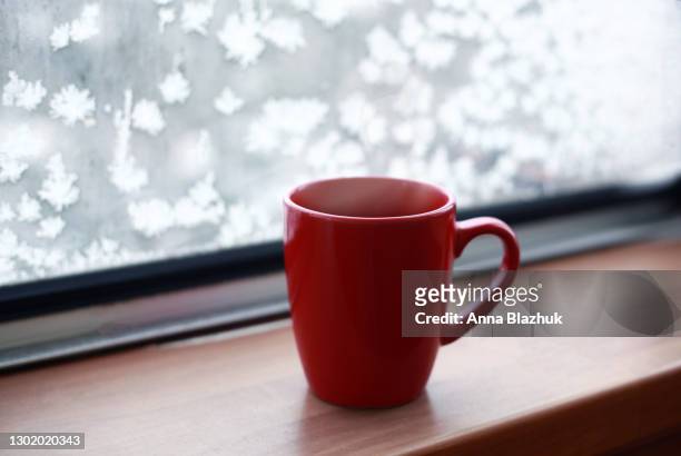 red cup of hot coffee against frozen window in the winter time. frost and cold weather concept. - window sill stock pictures, royalty-free photos & images