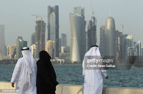 Men and women wearing traditional Qatari clothing visit the waterfront along the Persian Gulf across from new, budding financial district skyscrapers...