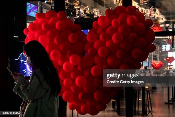 Women wear a mask while wear the mask uses iPhone in front of the heart shape balloon in Shopping Mall on February 14, 2021 in Wuhan, Hubei Province,...