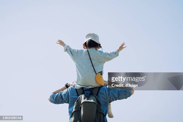 rear view of a loving young asian father carrying little daughter on his shoulders outdoors against sky. little daughter spreading her hands. they are spending some quality bonding time together on a lovey sunny day. family lifestyle - role reversal stock-fotos und bilder