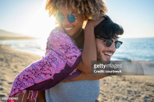 multi etniska par spelar piggyback på stranden vid solnedgången eller soluppgången. - young couple beach bildbanksfoton och bilder