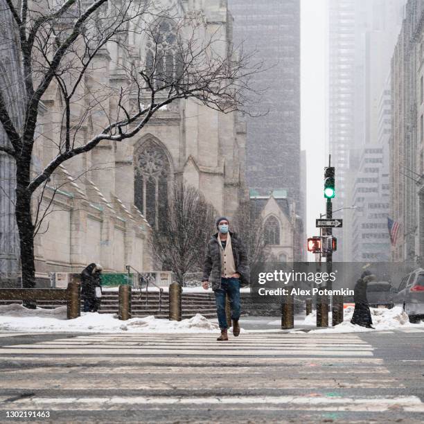 a young man wearing a protective mask crosses east 5th street along 5th avenue in manhattan in in front of st. patrick's cathedral during the winter storm and heavy snowfall. - alex potemkin coronavirus stock pictures, royalty-free photos & images