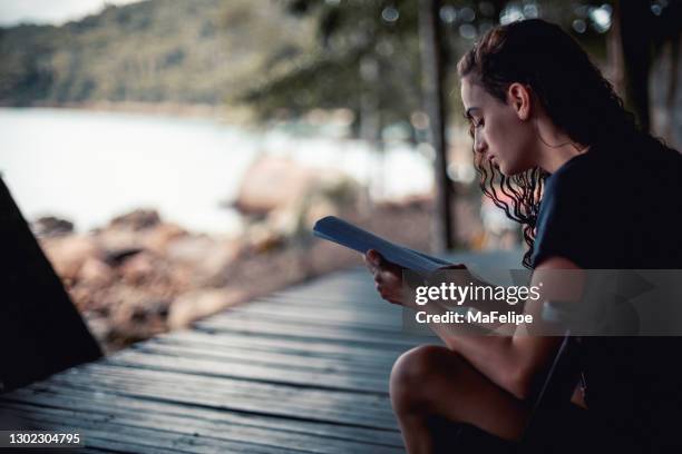 夕暮れ時にビーチのそばの木製バルコニーに座って本を読む若い女性 - angra dos reis ストックフォトと画像