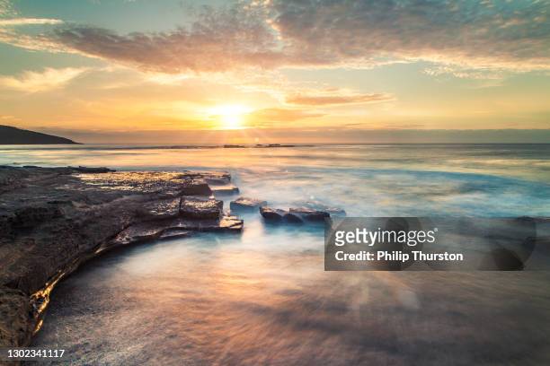 olas oceánicas que fluyen sobre rocas al amanecer con nubes de naranja y oro - vista marina fotografías e imágenes de stock