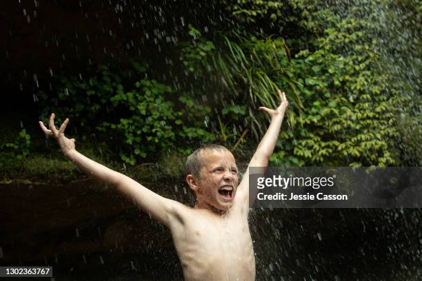 a boy stands topless under a waterfall with his arms in the air - drenched stock pictures, royalty-free photos & images