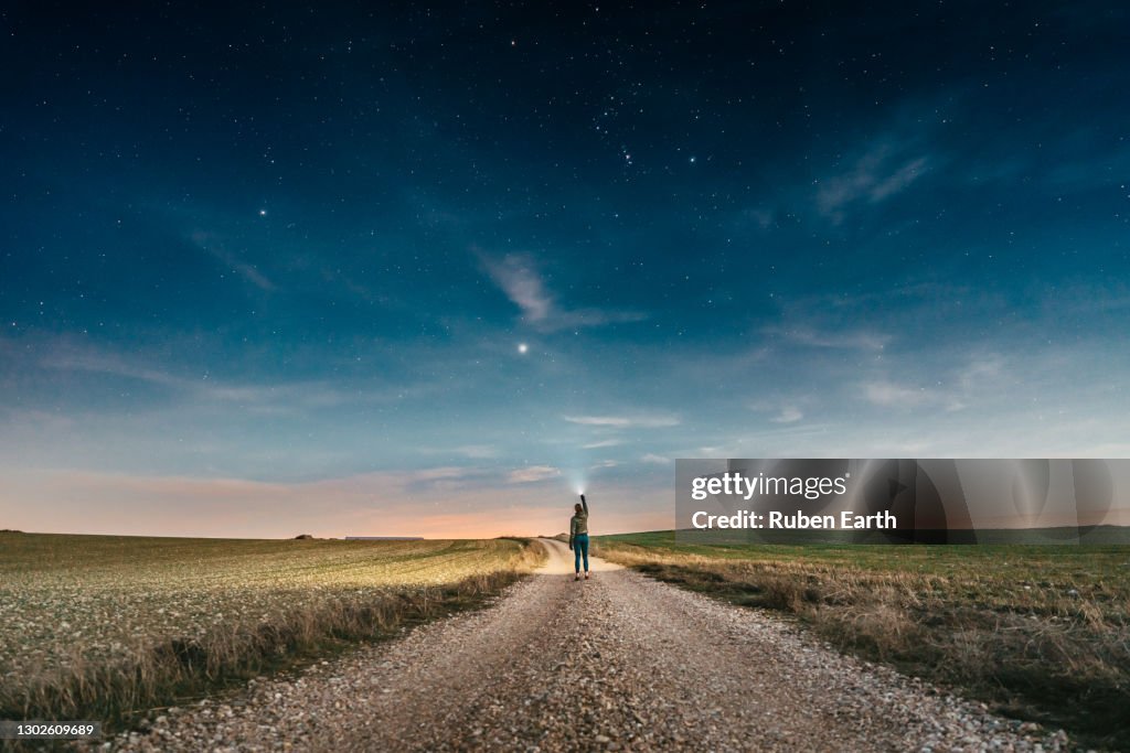 Woman pointing a lantern to the sky while walking on a country road at night with the clear sky full of stars