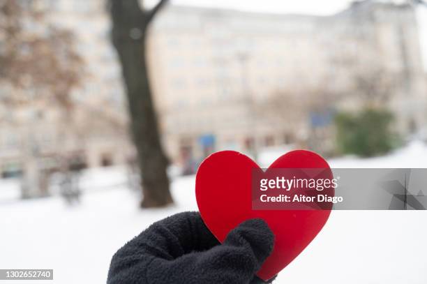 a person with winter globes holds a red heart shape while walking on the street in a snowy street in the city. berlin, germany. - frostbite fingers stock-fotos und bilder