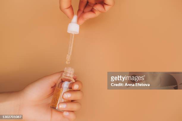 face serum in glass bottle with gold particles in women hand with beautiful nude colored manicure with gold particles and glass pipette in other hand on beige background. trendy colors of the year - nagelhaut stock-fotos und bilder