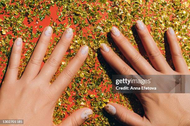 glowing hands with nude color manicure with gold particles above glittering red table. trendy colors of the year - gold nail polish stock-fotos und bilder