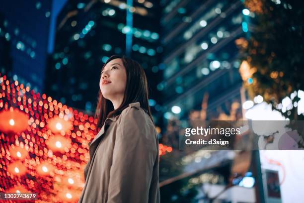 low angle portrait of young asian businesswoman standing in downtown city street, looking up with confidence against illuminated urban commercial buildings and vibrant city street lights at night. business on the go - chinese lantern festival stock-fotos und bilder