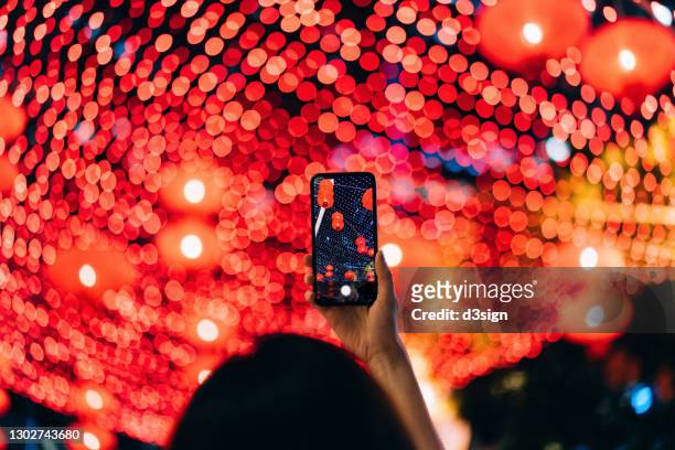 rear view of young asian woman taking photos of illuminated traditional chinese red lanterns with smartphone hanging along the city street at night. traditional chinese culture, festival and celebration event theme - hongkong lifestyle stock-fotos und bilder
