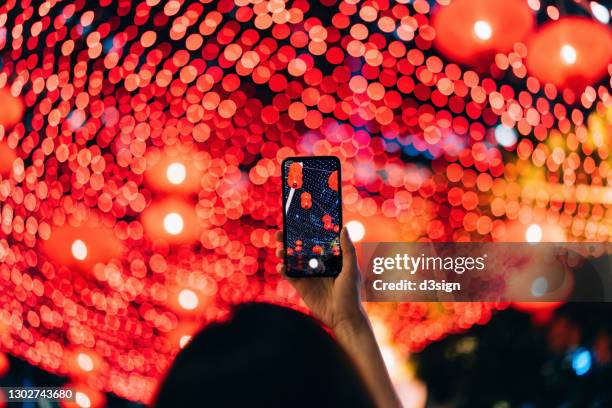 rear view of young asian woman taking photos of illuminated traditional chinese red lanterns with smartphone hanging along the city street at night. traditional chinese culture, festival and celebration event theme - hongkong lifestyle stockfoto's en -beelden