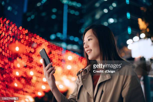low angle portrait of young asian businesswoman using smartphone in the city at night, against illuminated urban commercial buildings and vibrant city street lights. business on the go - chinese lantern festival stockfoto's en -beelden