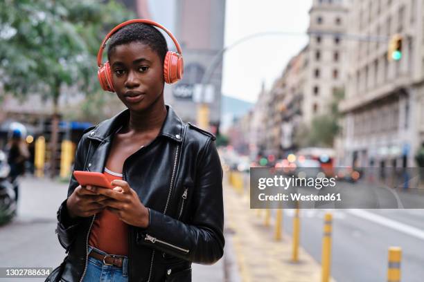 woman wearing headphones and jacket using smart phone while standing in city - transportation hearing stock-fotos und bilder