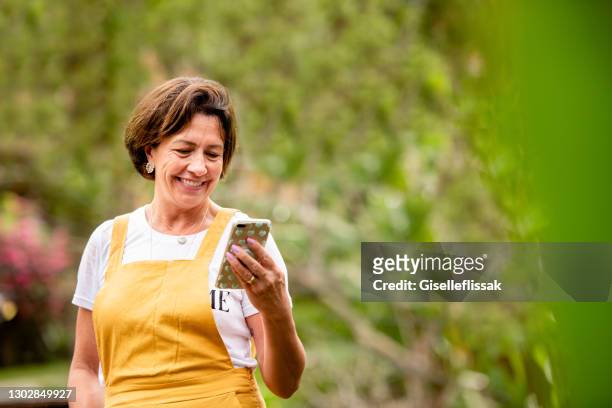 smiling mature woman video calling on a phone outside in her yard - mid adult women stock pictures, royalty-free photos & images