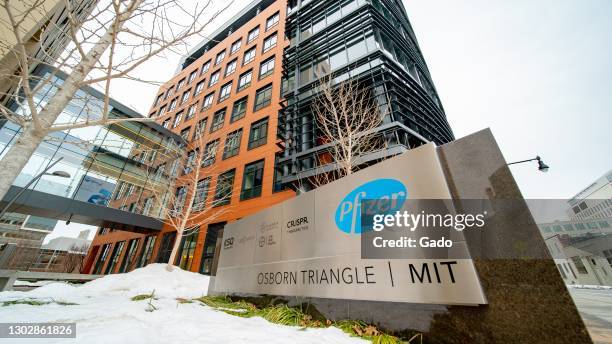 Low-angle view of Pfizer, MIT, CRISPR, KSQ, and other logos on a low sign, with buildings and glass bridge in the background and snow in the...